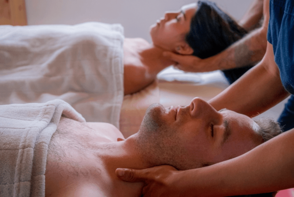 A couple relaxes under white towels as massage therapists gently work their necks and shoulders in a softly lit room, conveying calm and comfort.