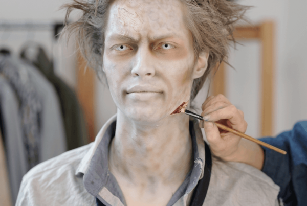 A pale-faced person with messy hair sits in costume as an unseen hand applies a bloody scratch to his neck, creating a tense zombie makeup scene.